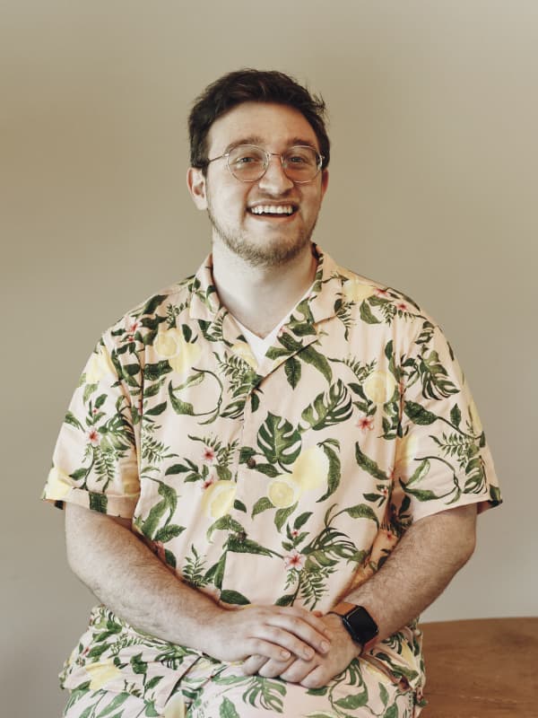 Josh in a full pink, Hawaiian lemon shirt and shorts outfit. His arms are crossed and he has a big smile.